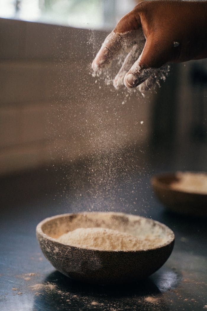 Close-up of a hand sprinkling flour into a ceramic bowl on a kitchen countertop.